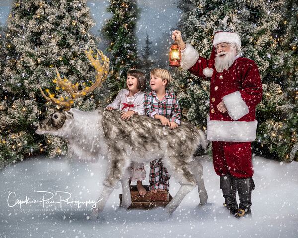 Two children with a reindeer while Santa Ray holds up a lantern in the snow