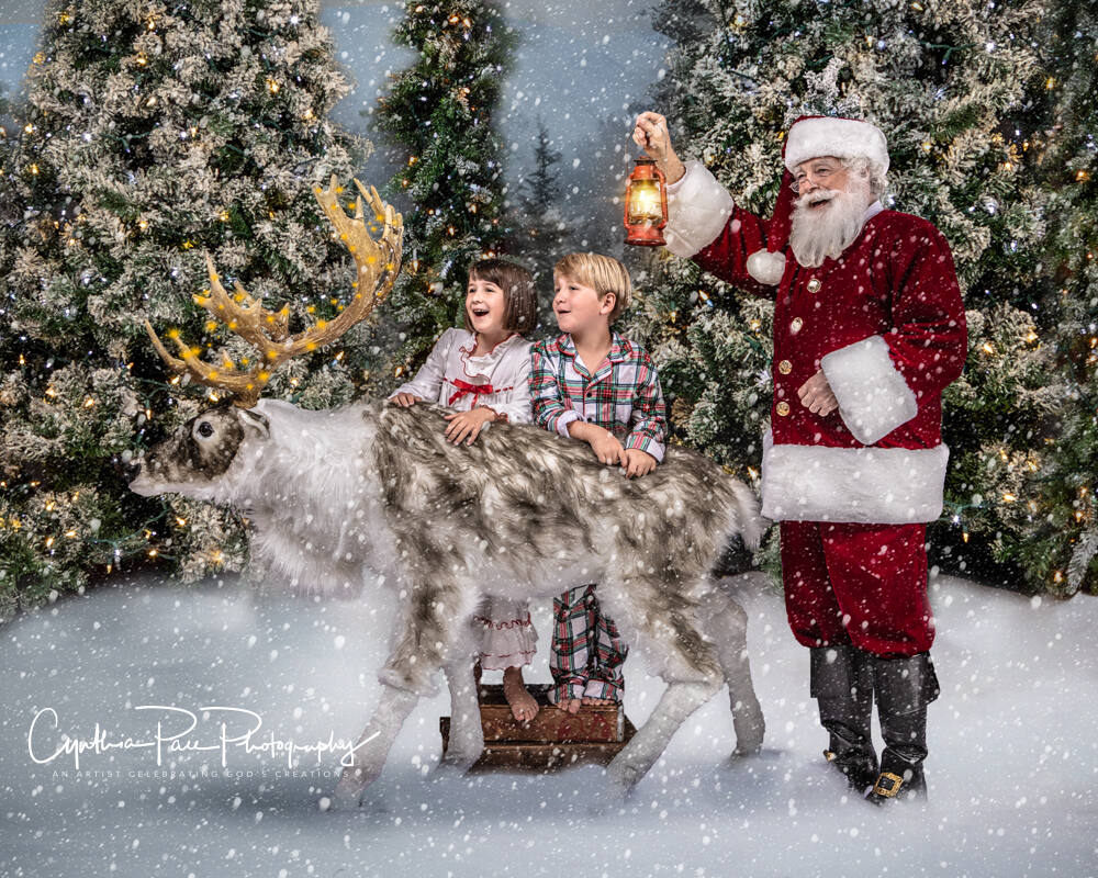 Two children with a reindeer while Santa Ray holds up a lantern in the snow